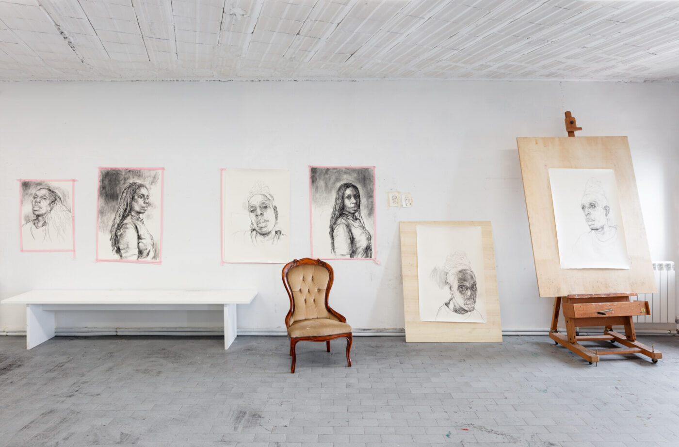 An art studio with portrait sketches on the walls and easels. A vintage chair sits in front of framed drawings on the floor. The room has a gray tile floor and a white ceiling with wooden beams.