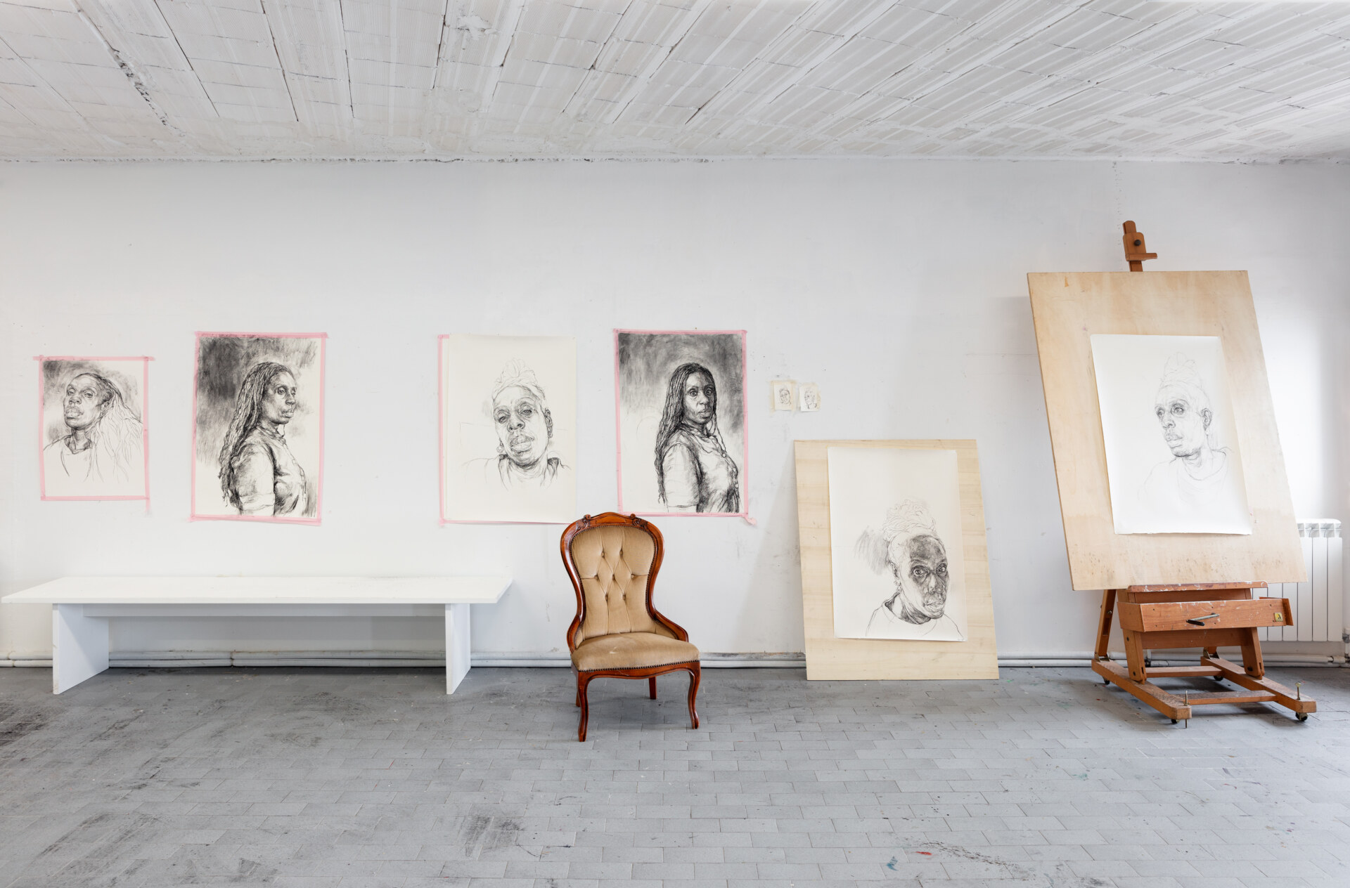 An art studio with portrait sketches on the walls and easels. A vintage chair sits in front of framed drawings on the floor. The room has a gray tile floor and a white ceiling with wooden beams.