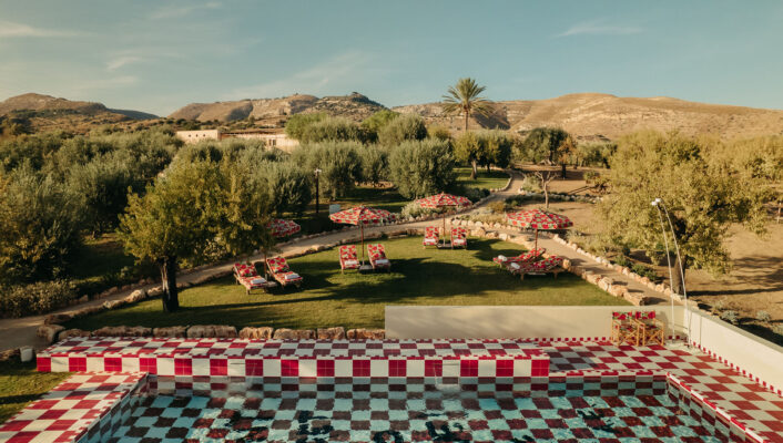A Tranquil Summer Retreat - A scenic view of a red and white checkered pool surrounded by sun loungers with floral cushions. Trees and a meandering pathway are in the background, and hills can be seen under a clear blue sky.
