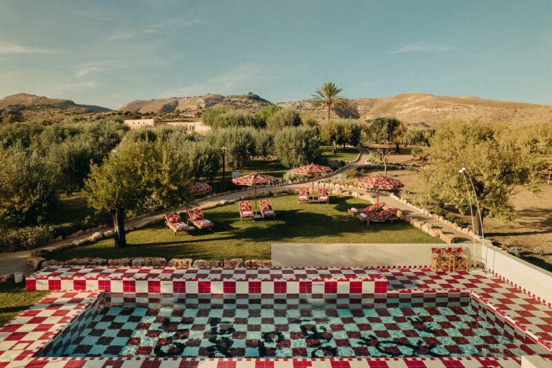 A Tranquil Summer Retreat - A scenic view of a red and white checkered pool surrounded by sun loungers with floral cushions. Trees and a meandering pathway are in the background, and hills can be seen under a clear blue sky.