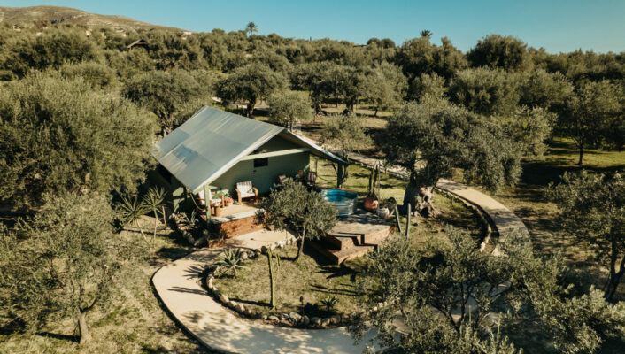 Aerial Retreat - Aerial view of a wooden cabin with a metal roof surrounded by trees and greenery. A circular path leads to the cabin, which features a patio and outdoor seating. The landscape is hilly with clear blue skies above.