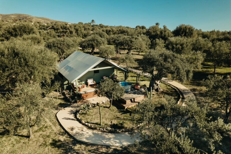 Aerial Retreat - Aerial view of a wooden cabin with a metal roof surrounded by trees and greenery. A circular path leads to the cabin, which features a patio and outdoor seating. The landscape is hilly with clear blue skies above.