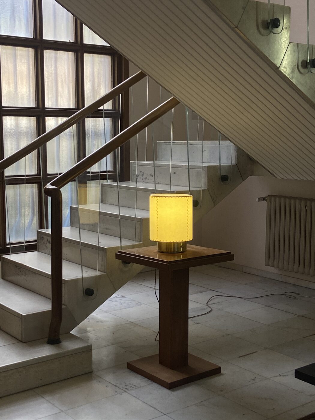 È visibile una scala moderna con ringhiera in legno e pannelli in vetro. Una lampada quadrata gialla è appoggiata su un piedistallo in legno di fronte alla scala. Il pavimento e le scale sono in pietra o piastrelle di colore chiaro.