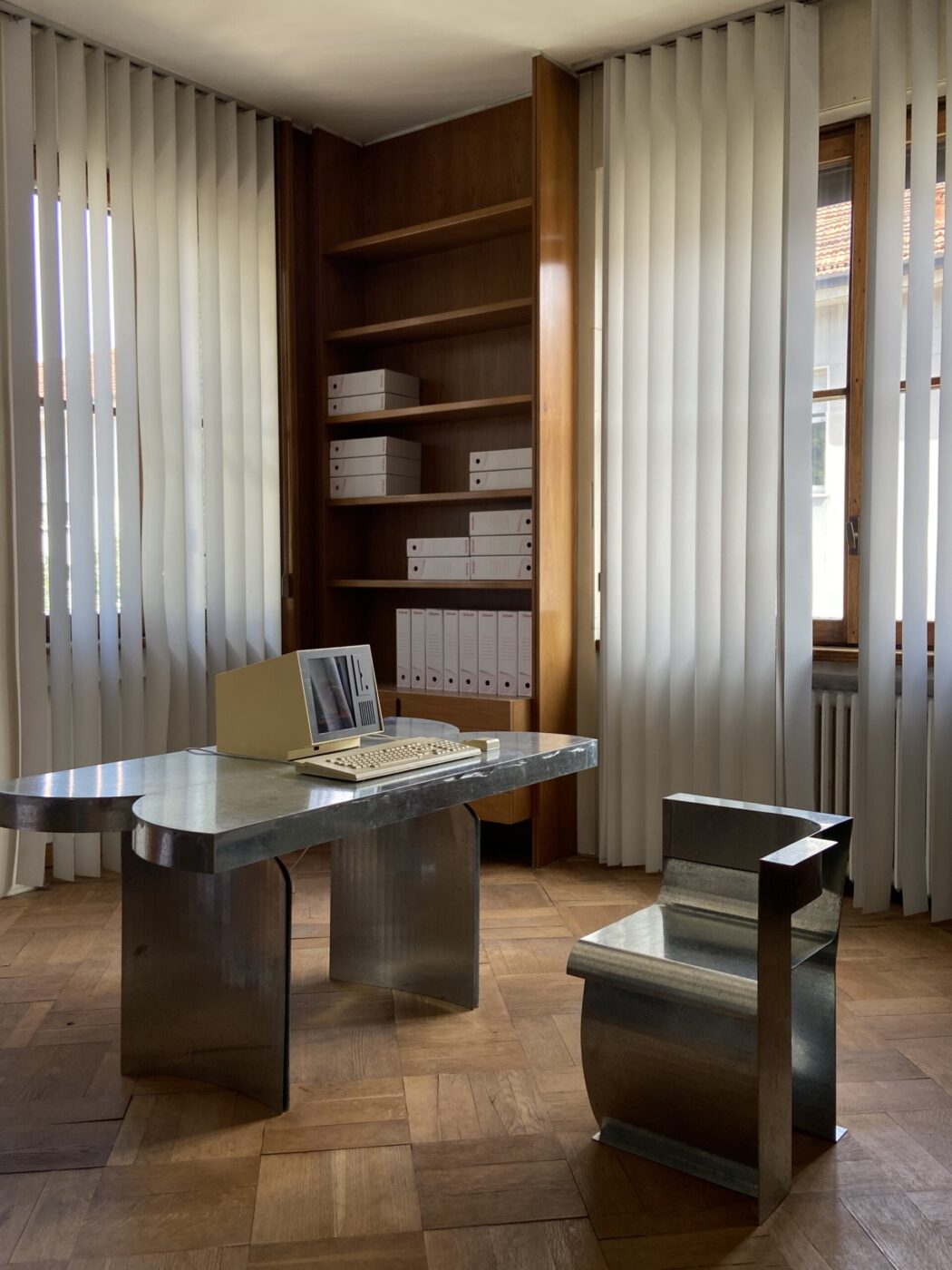 Vintage Workspace - Un ufficio minimalista con una scrivania e una sedia in metallo lucido. Un computer vintage è appoggiato sulla scrivania. Gli scaffali nell'angolo contengono raccoglitori bianchi ordinatamente organizzati. Le tende verticali coprono le finestre, diffondendo la luce naturale nella stanza.