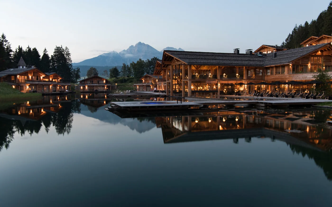 San Luis Lodges - A tranquil lakeside scene at dusk, featuring illuminated wooden cabins reflecting on the water. Mountains and trees form the backdrop, enhancing the serene atmosphere.