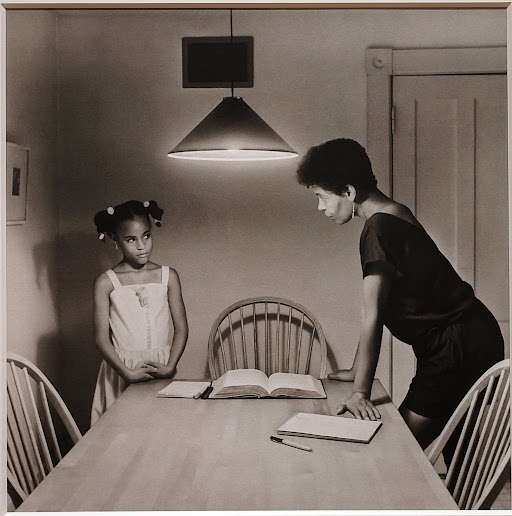 Conversation Around the Table - A black and white image of a woman leaning on a dining table, talking to a young girl in a dress standing on the opposite side. An open book and a notebook are on the table, lit by a hanging lamp. Chairs are arranged around the table.
