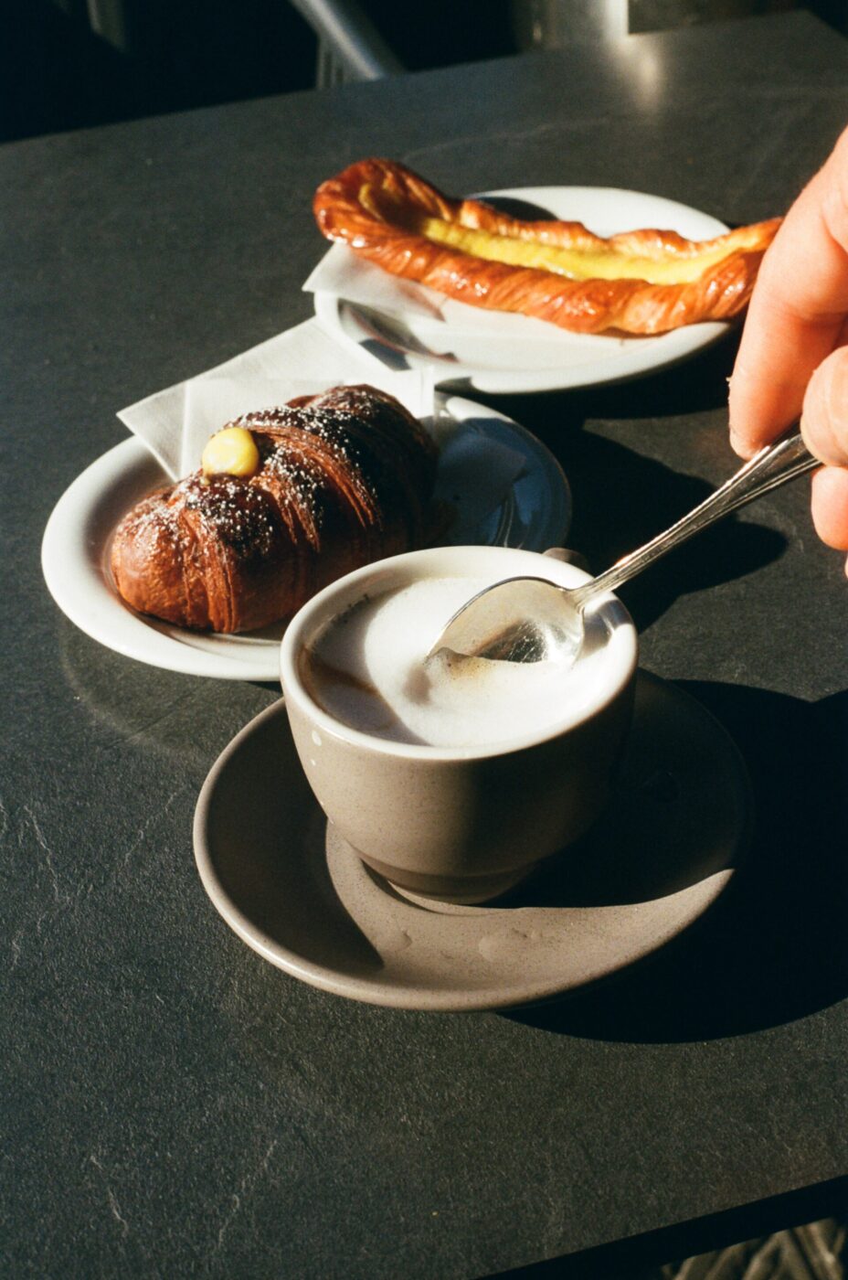 Morning Brew - A steaming cup of coffee sits on a saucer, with a hand stirring it using a spoon. In the background, a croissant topped with powdered sugar and a small pat of butter, and a twisted pastry on a plate rest on a dark surface in sunlight.