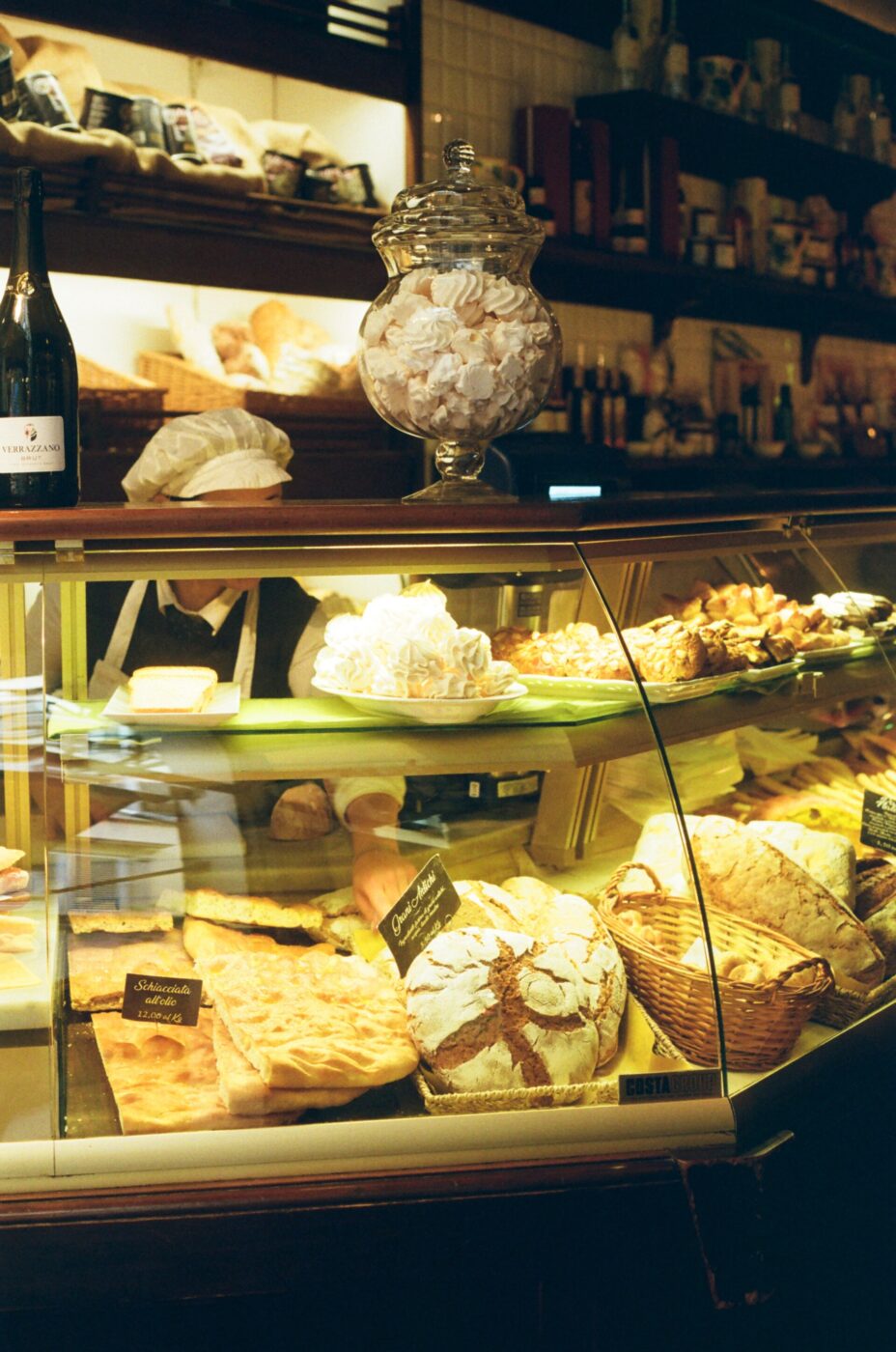 A bakery display case filled with various breads, pastries, and desserts. Items include loaves, cookies, and tarts. A large jar of meringues sits on top. Warm, ambient lighting creates a cozy, inviting atmosphere.