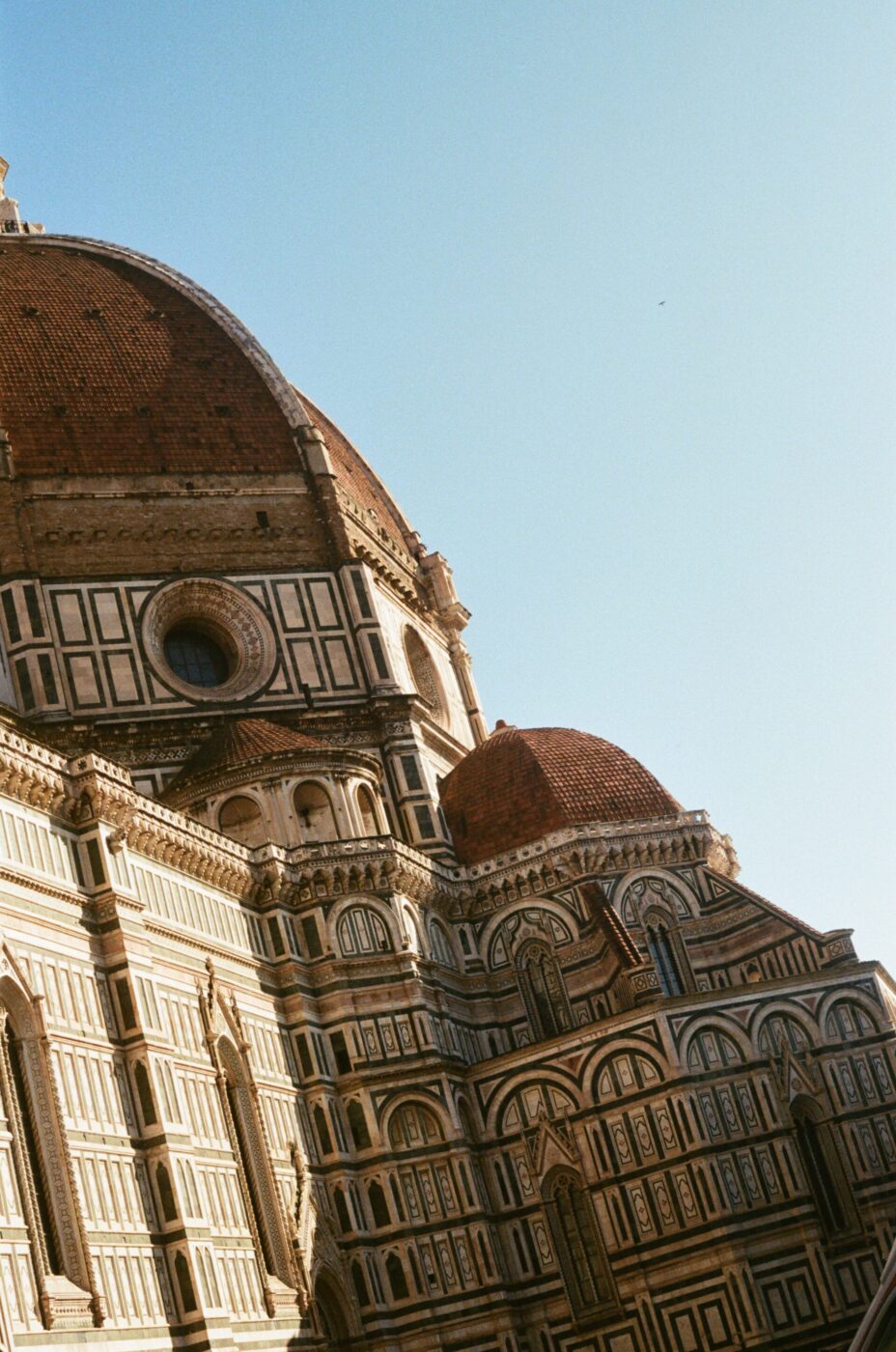 The Florence Cathedral with its iconic red-tiled dome and ornate facade, featuring intricate patterns and arched windows, against a clear blue sky.