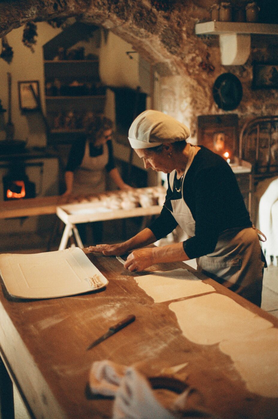 Cucina rustica" - Testo alternativo: Una donna con cuffia bianca e grembiule sta stendendo un impasto su un tavolo di legno in una cucina rustica. Un'altra persona sullo sfondo sta preparando gli ingredienti. La stanza è illuminata da una luce calda e si vede un forno tradizionale.