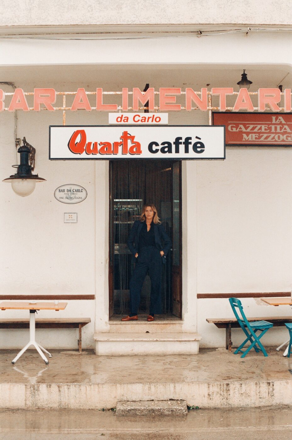 Quarta caffè e Bar Alimentari da Carlo" - Una persona è in piedi sulla soglia di un edificio sotto un'insegna con la scritta "Quarta caffè e Bar Alimentari da Carlo". Fuori, in una giornata piovosa, ci sono tavoli e sedie, con un'insegna al neon rosa sopra.