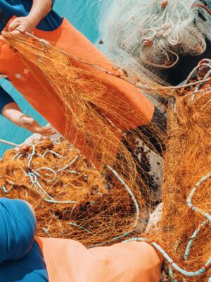 Salento - Fishers wearing blue shirts and orange pants handle large, orange fishing nets on a boat. The turquoise sea is visible in the background, with the nets and ropes spread out, suggesting an active fishing scene.