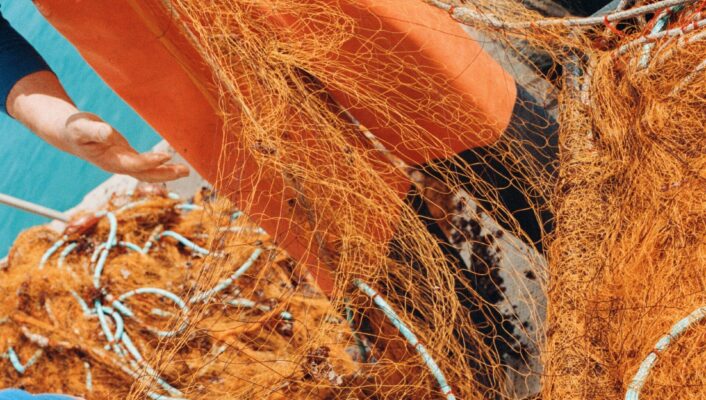 Salento - Fishers wearing blue shirts and orange pants handle large, orange fishing nets on a boat. The turquoise sea is visible in the background, with the nets and ropes spread out, suggesting an active fishing scene.