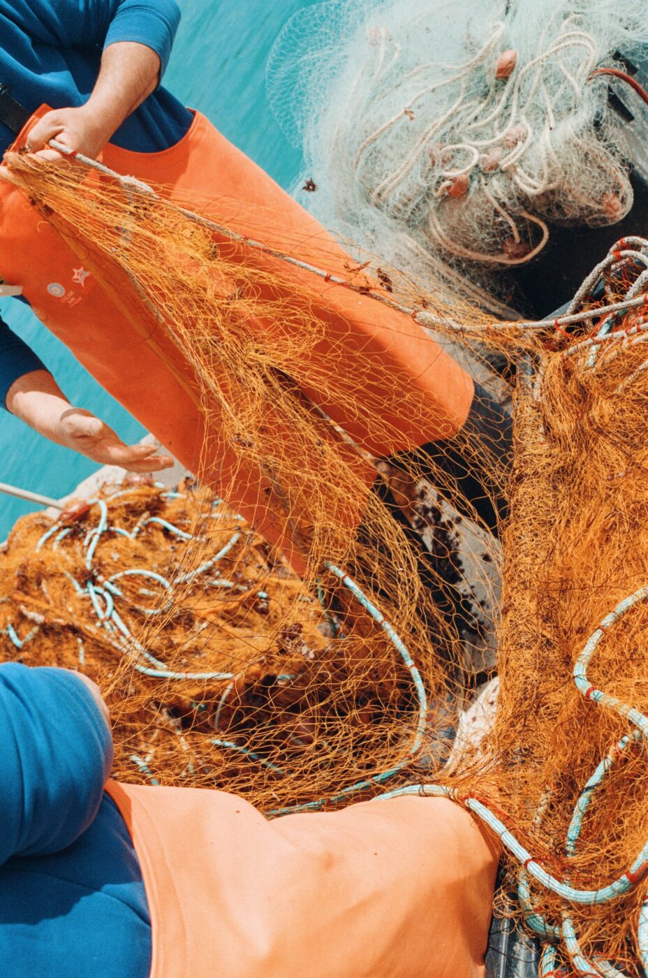 Due pescatori in mare - Due pescatori in tuta arancione maneggiano reti da pesca arancioni aggrovigliate su una barca. Sullo sfondo si vede il mare blu. La scena raffigura una tipica giornata di lavoro in mare.