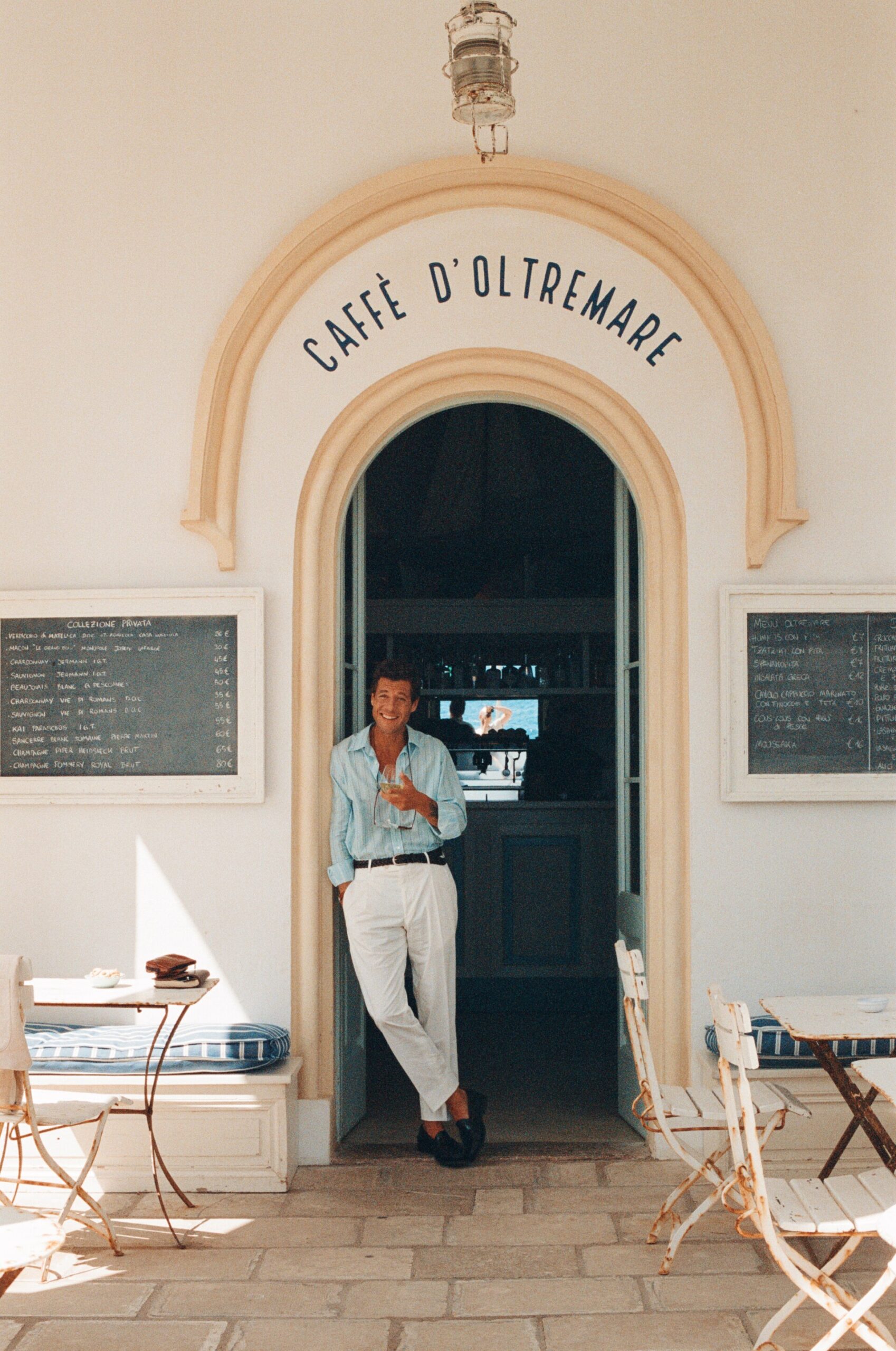Ingresso del Caffè D'Oltremare - Una persona in camicia blu e pantaloni bianchi si appoggia con nonchalance sulla soglia del Caffè D'Oltremare. Il nome del caffè è esposto in alto, con i menu ai lati. Sedie e tavoli vuoti sono in primo piano, sotto una luce soffusa esterna.
