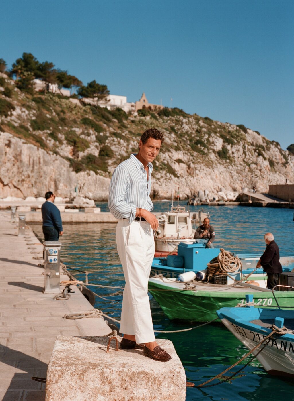 Momento di gioia nel porto - Un uomo con una camicia a righe blu e bianche e pantaloni bianchi è in piedi su un molo in riva al mare, sorridente. Barche e pescatori sono visibili sullo sfondo. Alle loro spalle si erge una collina rocciosa con cespugli verdi sotto un cielo azzurro e terso.