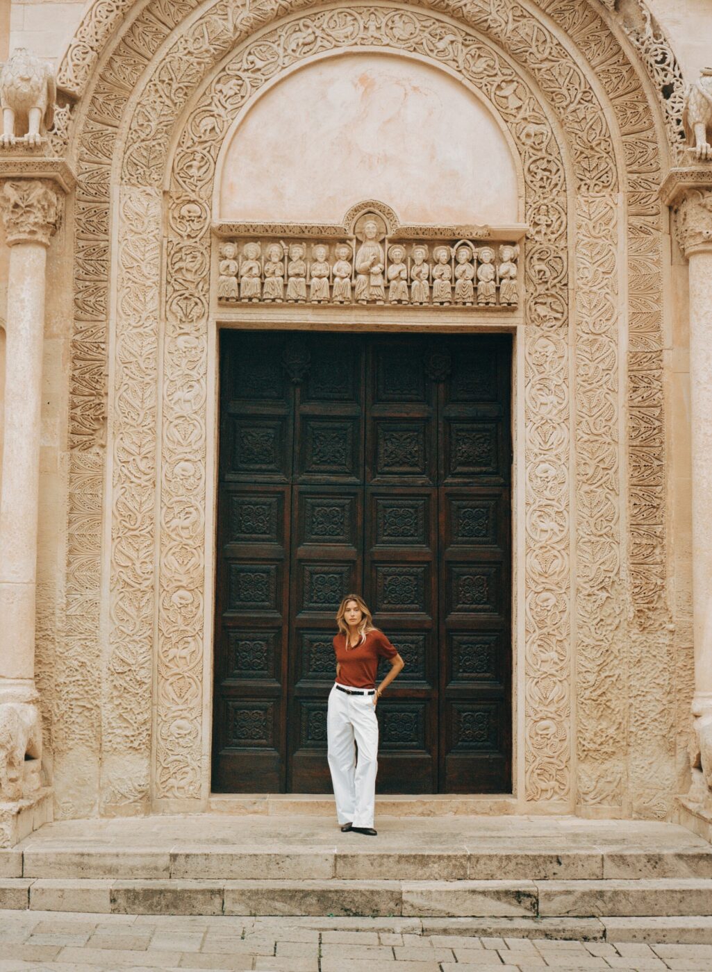 [Titolo dell'immagine] - [Testo alternativo]: Una persona è in piedi davanti a una porta di legno ad arco decorata, con indosso una maglietta rossa e pantaloni bianchi. La facciata in pietra presenta intricati intagli e colonne.