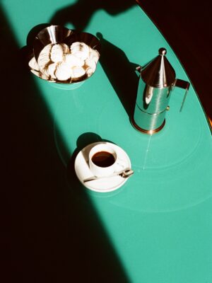 Conica Espresso Coffee Maker : Teal Table with Meringue Cookies and Espresso - A teal table with a bowl of meringue cookies, a silver coffee maker, and a cup of espresso on a saucer with a spoon. Strong sunlight casts dramatic shadows across the scene.