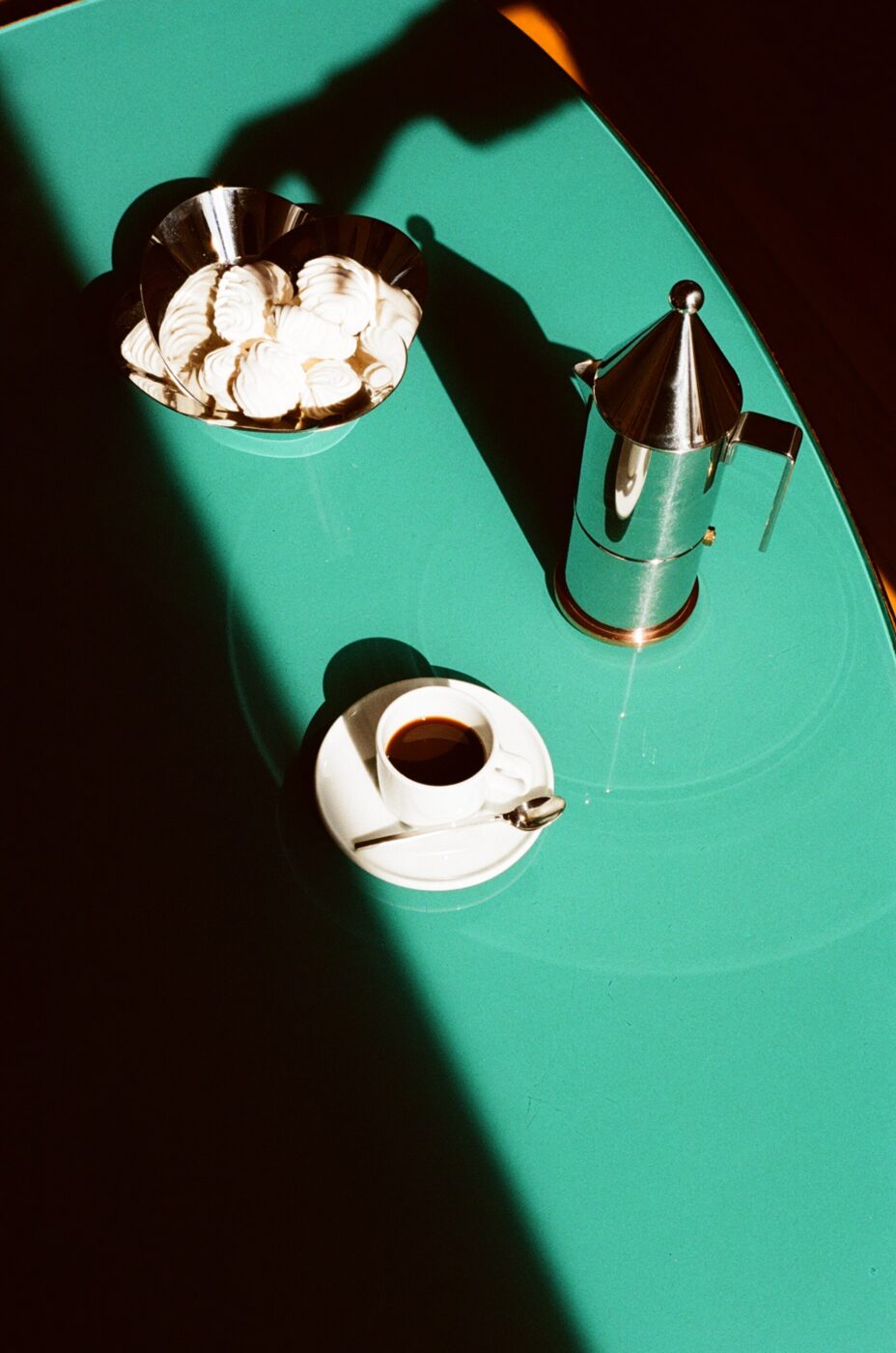 Conica Espresso Coffee Maker : Teal Table with Meringue Cookies and Espresso - A teal table with a bowl of meringue cookies, a silver coffee maker, and a cup of espresso on a saucer with a spoon. Strong sunlight casts dramatic shadows across the scene.