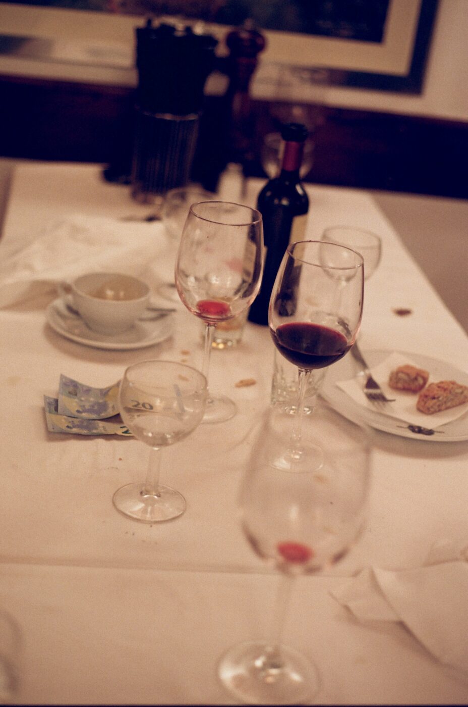 Trattoria Cammillo - A dimly lit table with several empty wine glasses, a wine bottle, a teacup with saucer, a few banknotes, and a plate of partially eaten food on a white tablecloth. A pepper grinder is visible in the background.