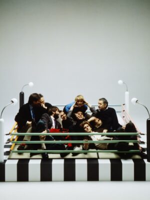 Zebra-Striped Boxing Ring Meeting - A group of people sit closely together inside a small, zebra-striped boxing ring with colorful ropes, surrounded by four white corner poles, each topped with a light. The setting is minimalistic, with a plain, neutral background.