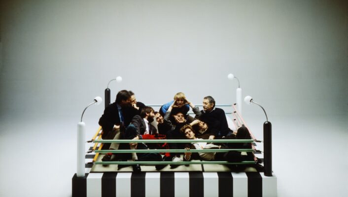 Zebra-Striped Boxing Ring Meeting - A group of people sit closely together inside a small, zebra-striped boxing ring with colorful ropes, surrounded by four white corner poles, each topped with a light. The setting is minimalistic, with a plain, neutral background.