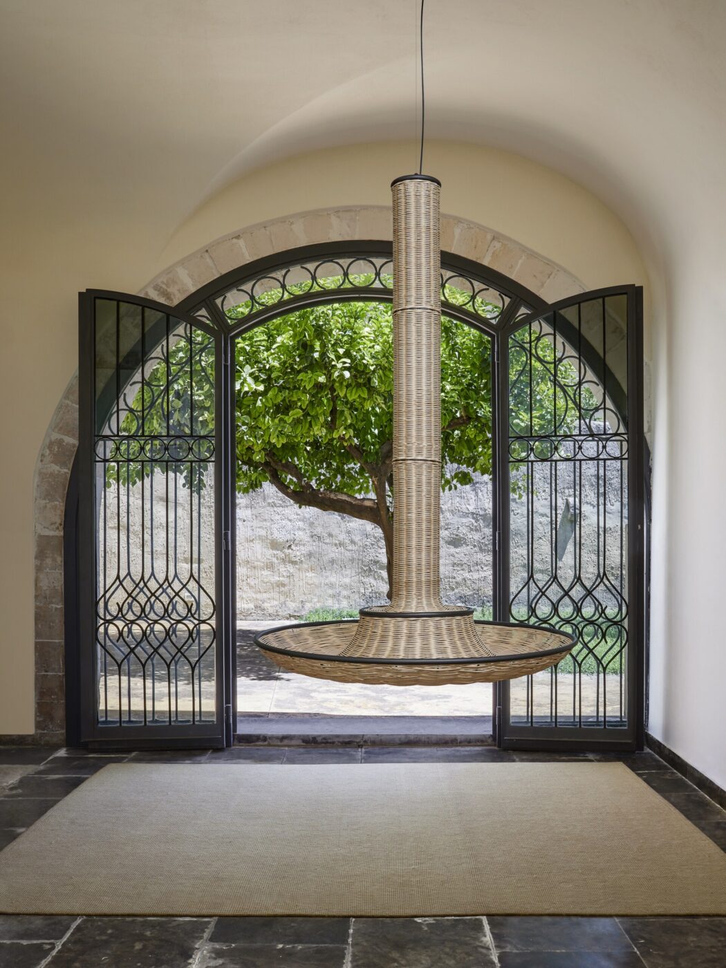 Non è stato fornito alcun titolo per l'immagine, quindi il testo alternativo dovrebbe essere: Una moderna sedia sospesa intrecciata con uno schienale alto è sospesa davanti a grandi porte ad arco doppie che si aprono su un giardino con un albero frondoso e un muro di pietra, riempiendo lo spazio di luce naturale.
