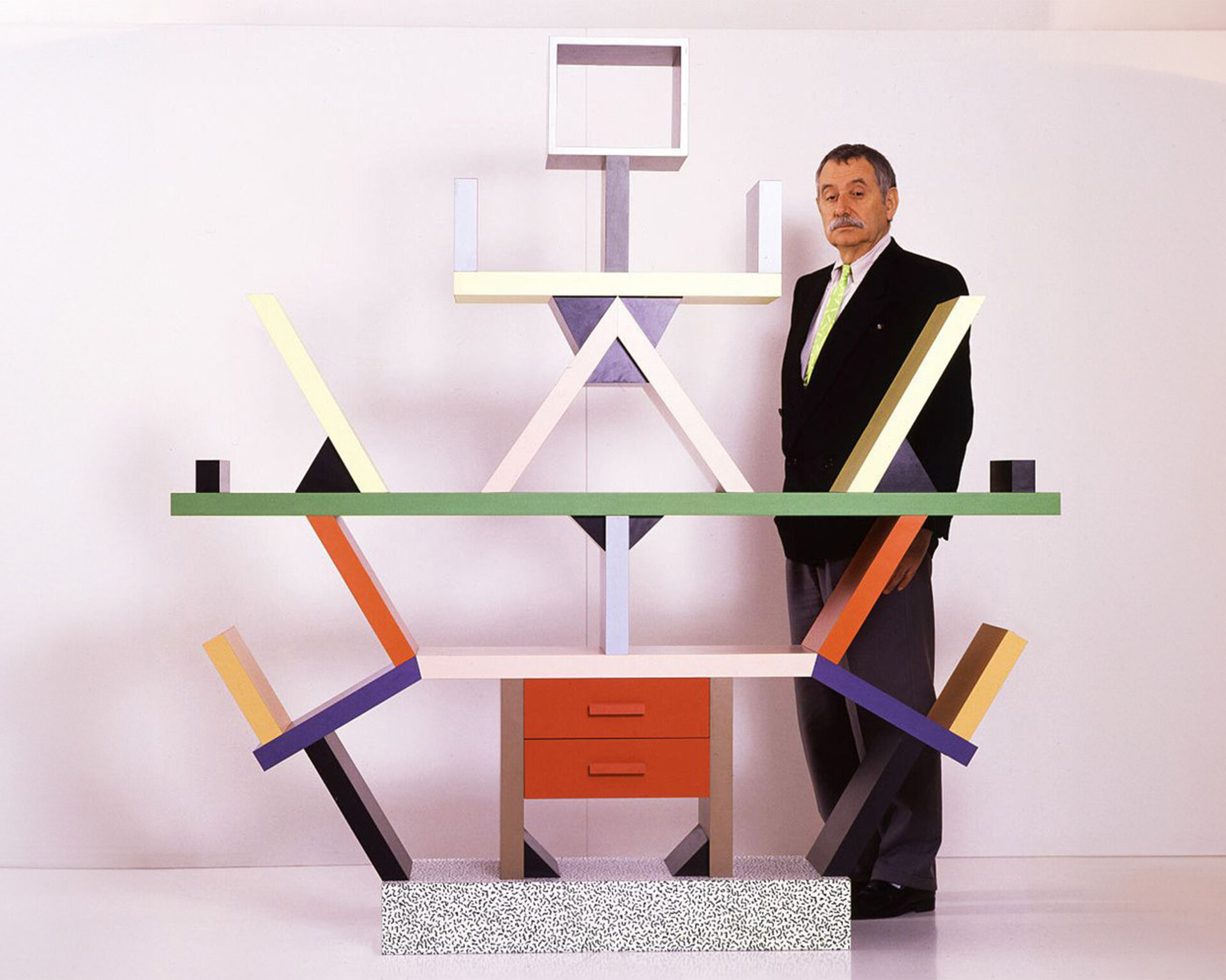 Geometric Wonder - Una persona è in piedi accanto a una libreria geometrica e colorata con più scomparti e linee angolari. La struttura include colori audaci come il verde, il viola e il giallo, con una base punteggiata e un cassetto rosso al centro.