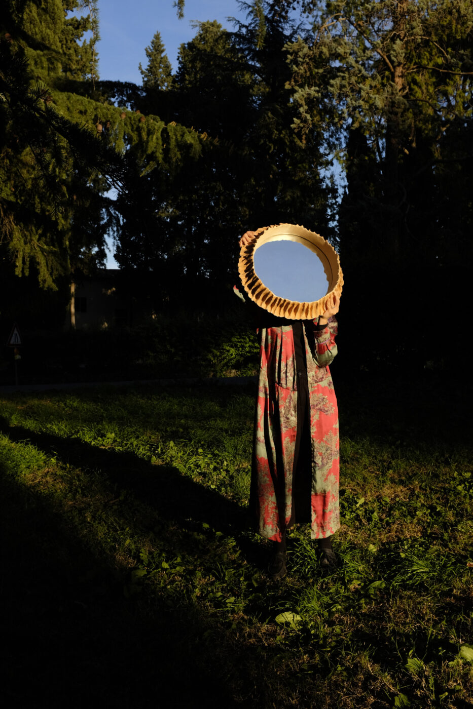 [Riflesso di uno specchio rotondo] - Una persona con un lungo abito a fantasia è in piedi sull'erba e tiene uno specchio rotondo davanti al viso, che riflette il cielo azzurro e gli alberi. La scena è ambientata all'aperto, con alberi alti e la luce del sole che crea ombre profonde.