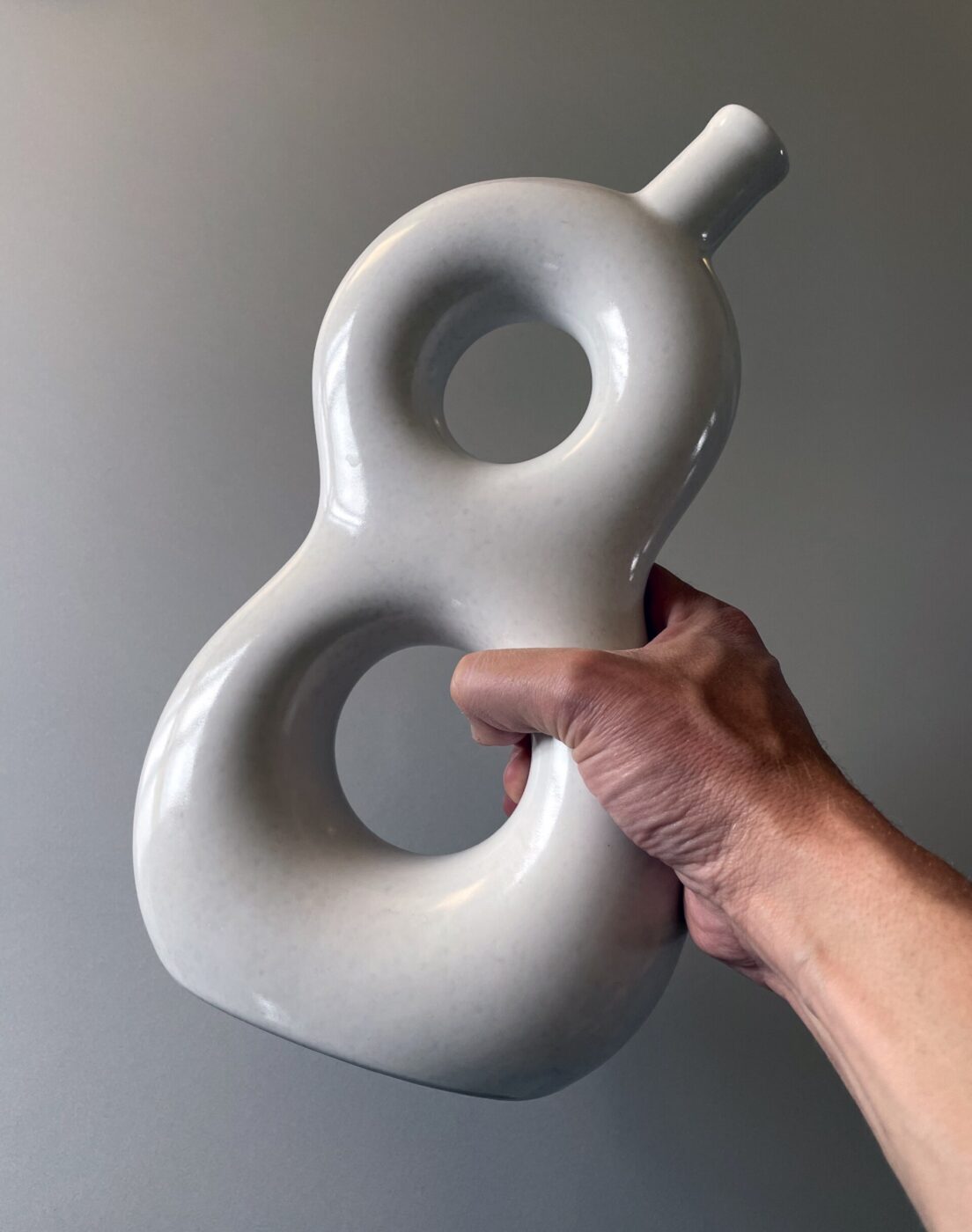 Una mano che regge un vaso liscio di ceramica bianca con due grandi aperture circolari, una sopra l'altra, che ricordano il numero otto su uno sfondo grigio semplice.