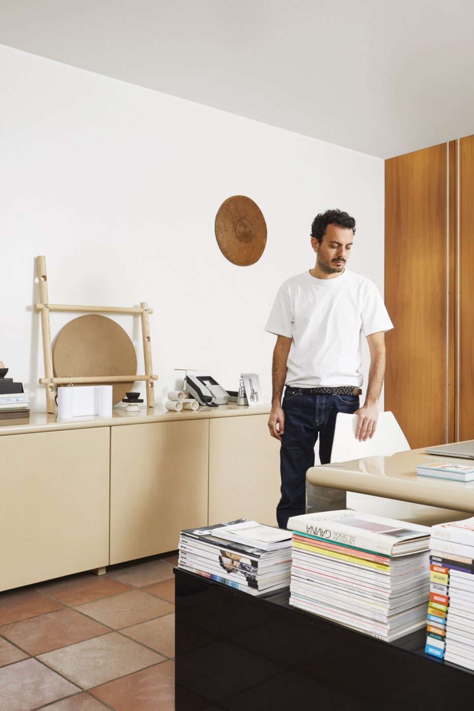 Certo! Ecco il testo alternativo formattato, supponendo che ci sia un titolo per l'immagine (userò "Ritratto di un ufficio moderno" come segnaposto; sostituisci il titolo effettivo se ne hai uno): Ritratto di un ufficio moderno - Un uomo con una maglietta bianca è in piedi in un ufficio moderno con un mobiletto beige, pile di riviste su un tavolo nero e un arredamento minimalista, inclusi oggetti in legno alla parete. Se fornisci il titolo specifico dell'immagine, posso aggiornarlo di conseguenza.
