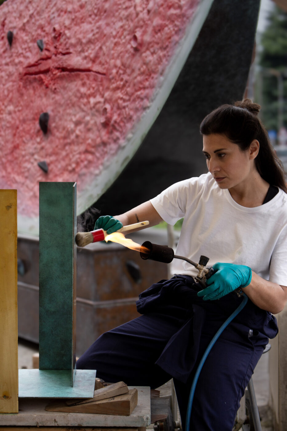 Una donna che indossa i guanti utilizza una fiamma ossidrica e una spazzola per lavorare su una scultura in metallo all'aperto, con una grande scultura a forma di anguria sullo sfondo.