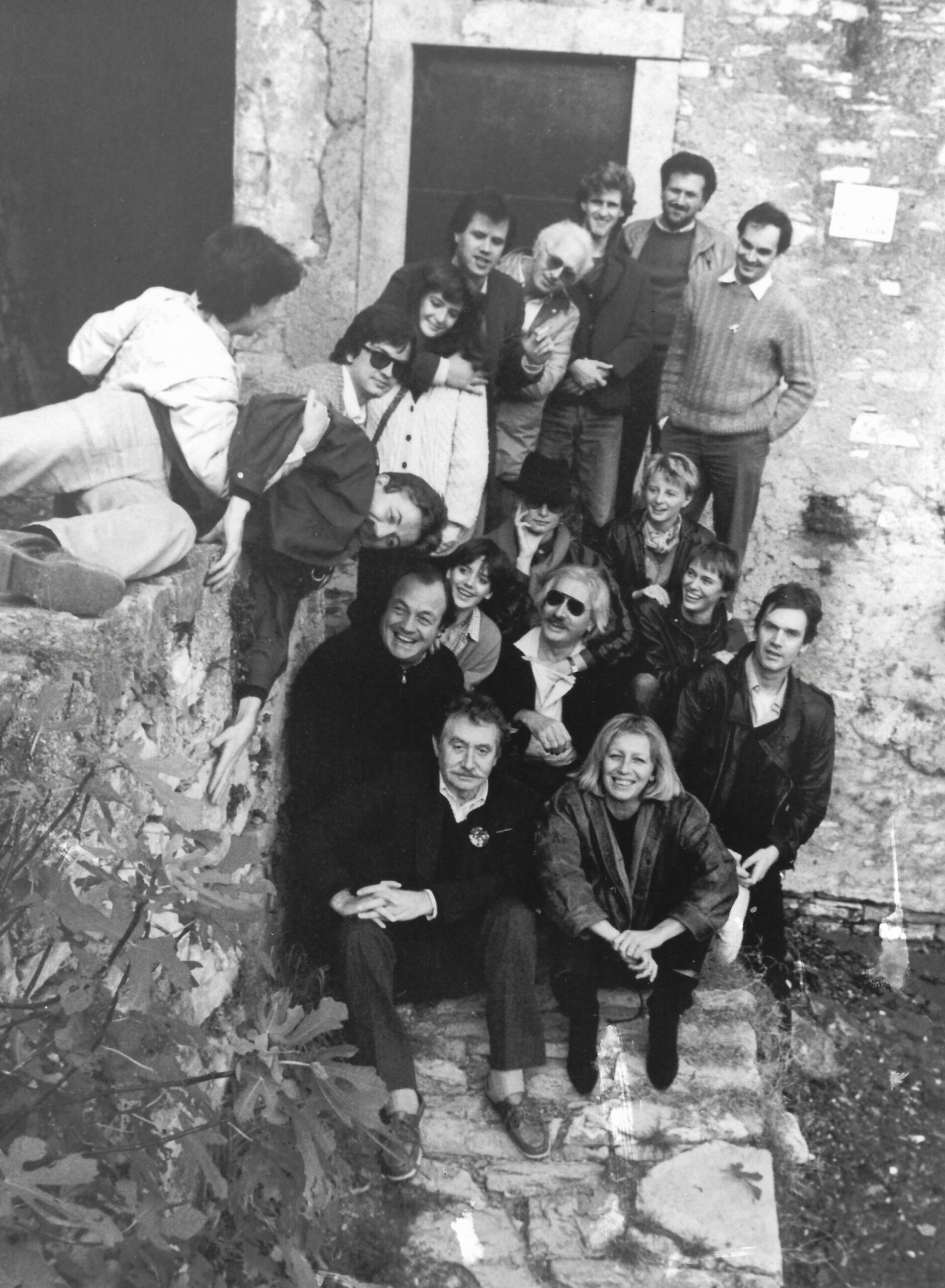 Raduno all'aperto" - Un gruppo di persone, vestite in modo casual, si riunisce all'esterno su una scalinata in pietra in una foto in bianco e nero. Alcuni sono seduti, altri in piedi o appoggiati, tutti sorridenti o dall'aspetto rilassato in un ambiente rustico all'aperto.