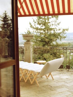 Un accogliente rifugio nel patio - Un accogliente patio con una poltrona imbottita e un tavolino si trova sotto una tenda a righe rosse e bianche, con vista panoramica sugli alberi e su una valle lontana. Il pavimento in pietra e una ringhiera in ferro battuto completano l'ambiente.