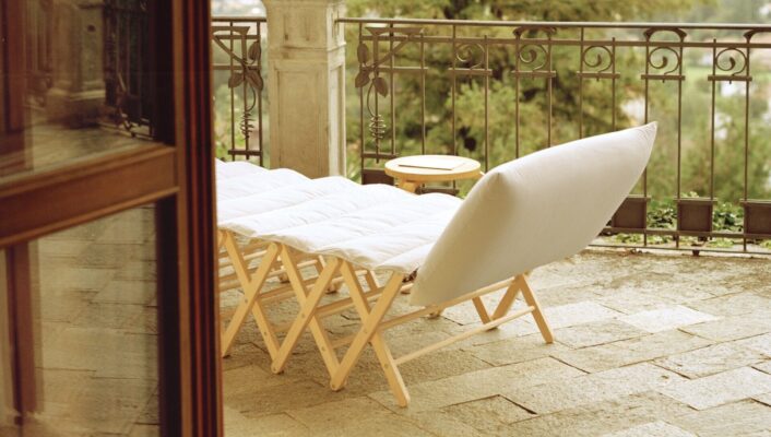 A cushioned lounge chair and small table sit on a stone patio under a red-and-white striped awning, overlooking a scenic landscape with trees and a distant view.