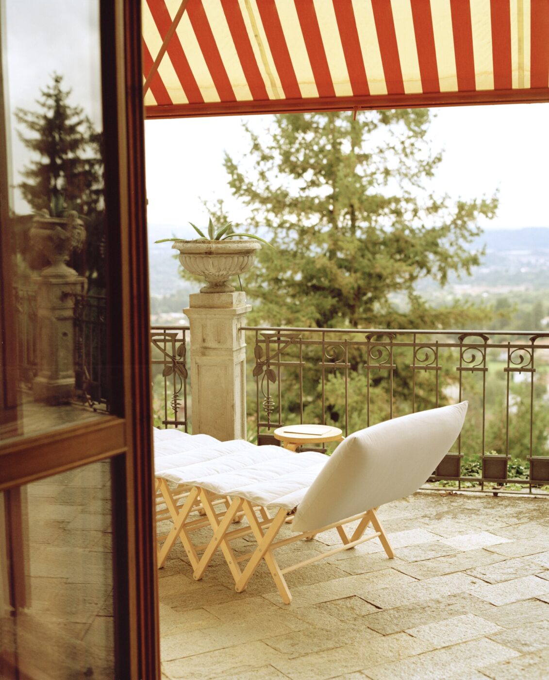 [Nessun titolo dell'immagine] - Una poltrona imbottita e un tavolino sono sistemati su un patio in pietra sotto una tenda a strisce rosse e bianche, con vista su un panorama con alberi e una ringhiera decorata.