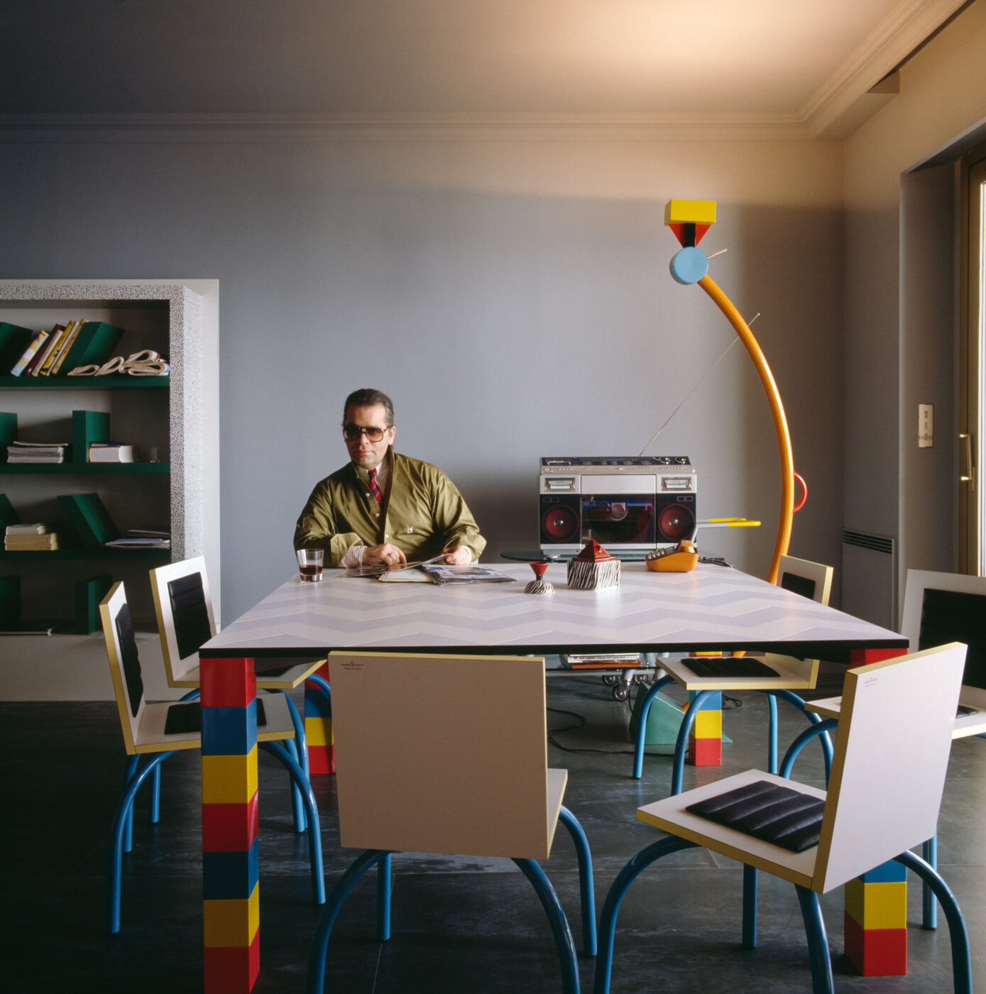 Una persona seduta a un tavolo da pranzo colorato e geometrico con sedie abbinate. Sullo sfondo c'è una libreria e sul tavolo c'è uno stereo retrò. La stanza ha un'atmosfera moderna e artistica con una lampada da terra unica e colorata.