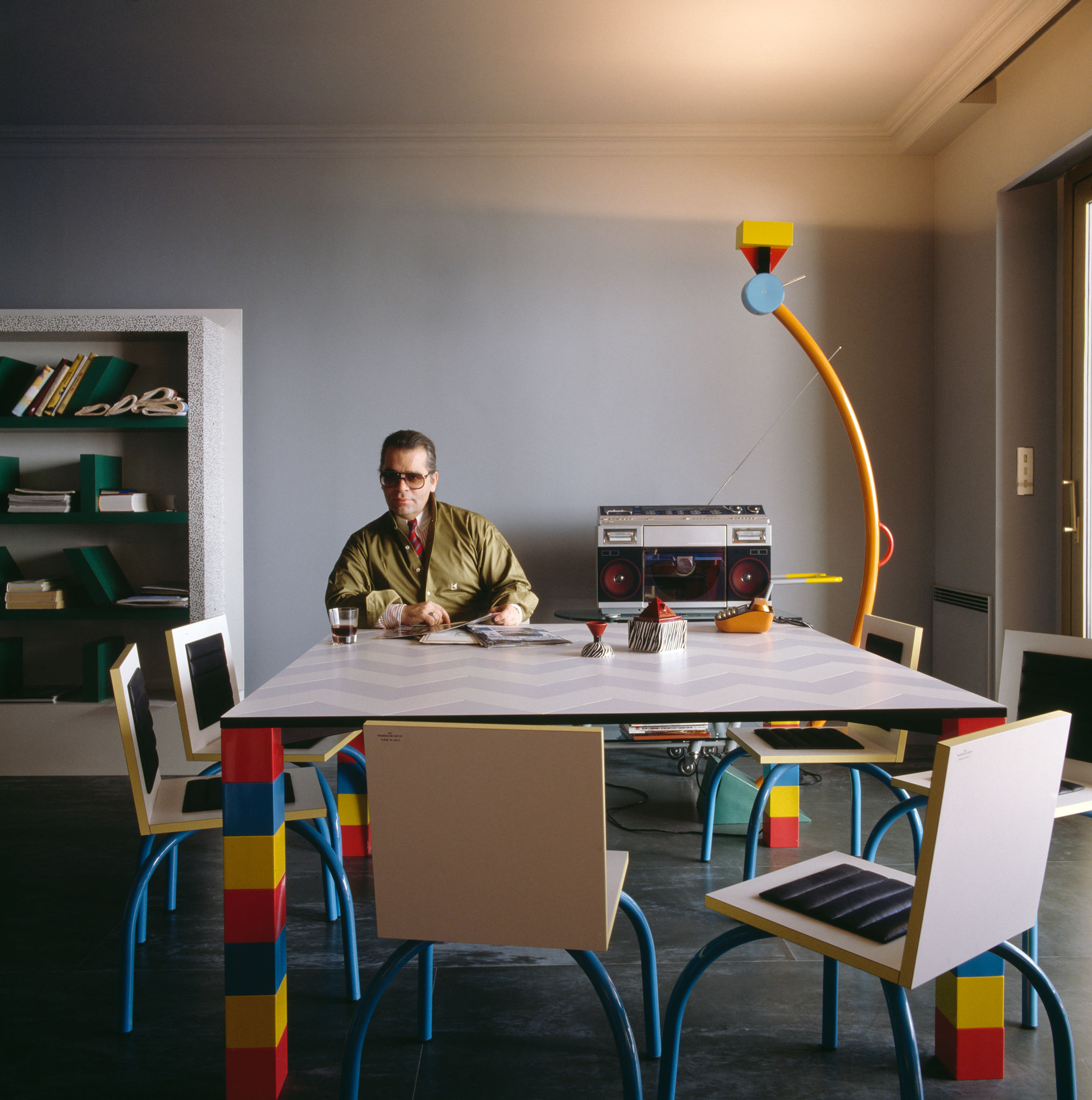 Una persona seduta a un tavolo da pranzo colorato e geometrico con sedie abbinate. Sullo sfondo c'è una libreria e sul tavolo c'è uno stereo retrò. La stanza ha un'atmosfera moderna e artistica con una lampada da terra unica e colorata.