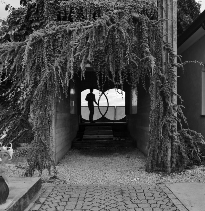 Tomba Brion: A silhouette of a person standing in the distance framed by a circular window. The pathway leading to the window is flanked by lush, hanging vines and framed by architectural elements, creating a mysterious atmosphere.