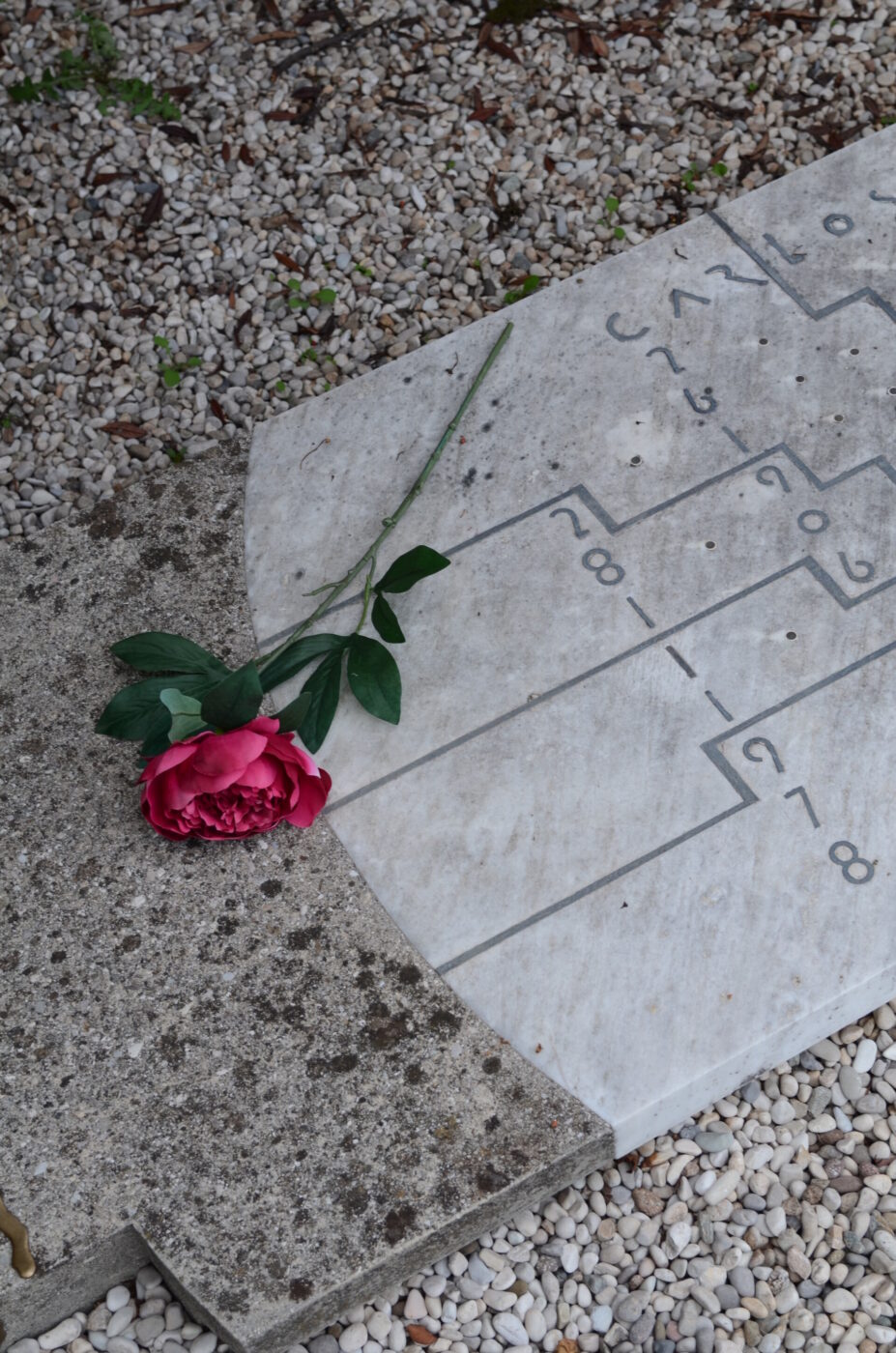 [Titolo dell'immagine] - Una lapide di marmo su una superficie di ghiaia con una rosa rosa adagiata su di essa. La pietra ha linee e numeri incisi, che ricordano un diagramma o un diagramma di flusso. La scena evoca un senso di ricordo e tranquillità.