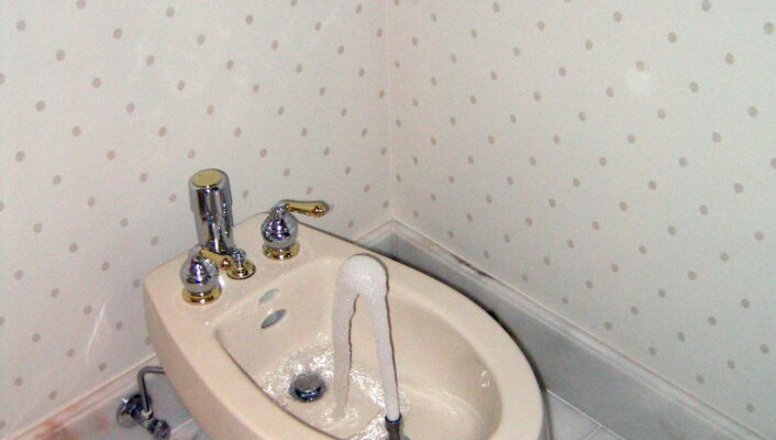 Classic Bathroom Bidet - A beige bidet with water flowing from its central spout is installed in a bathroom with patterned wallpaper and marble tile flooring. Two white towels hang on the wall above it. If you provide a different image title, simply replace Classic Bathroom Bidet with your provided title at the beginning of the description.