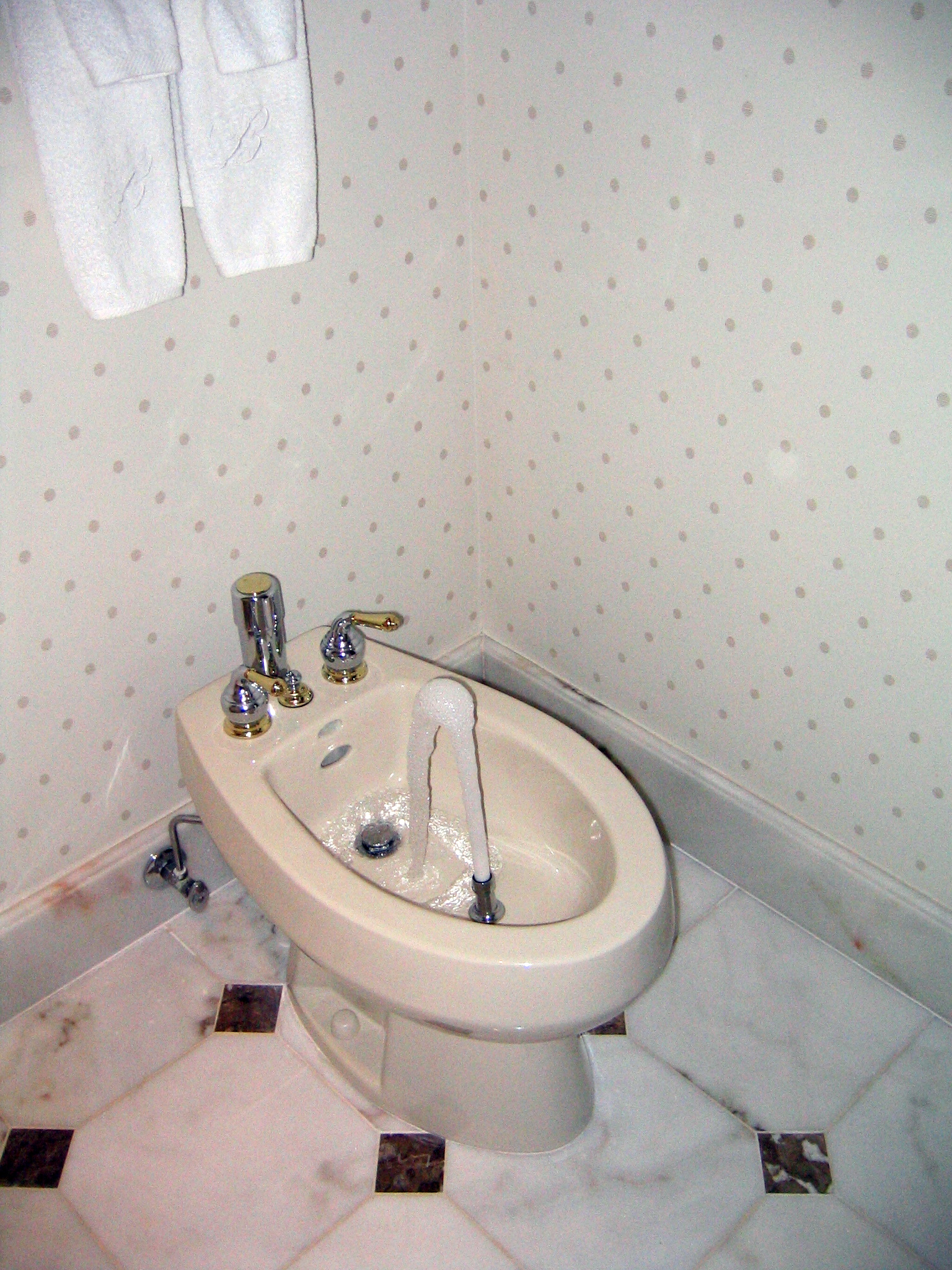 Classic Bathroom Bidet - A beige bidet with water flowing from its central spout is installed in a bathroom with patterned wallpaper and marble tile flooring. Two white towels hang on the wall above it. If you provide a different image title, simply replace Classic Bathroom Bidet with your provided title at the beginning of the description.
