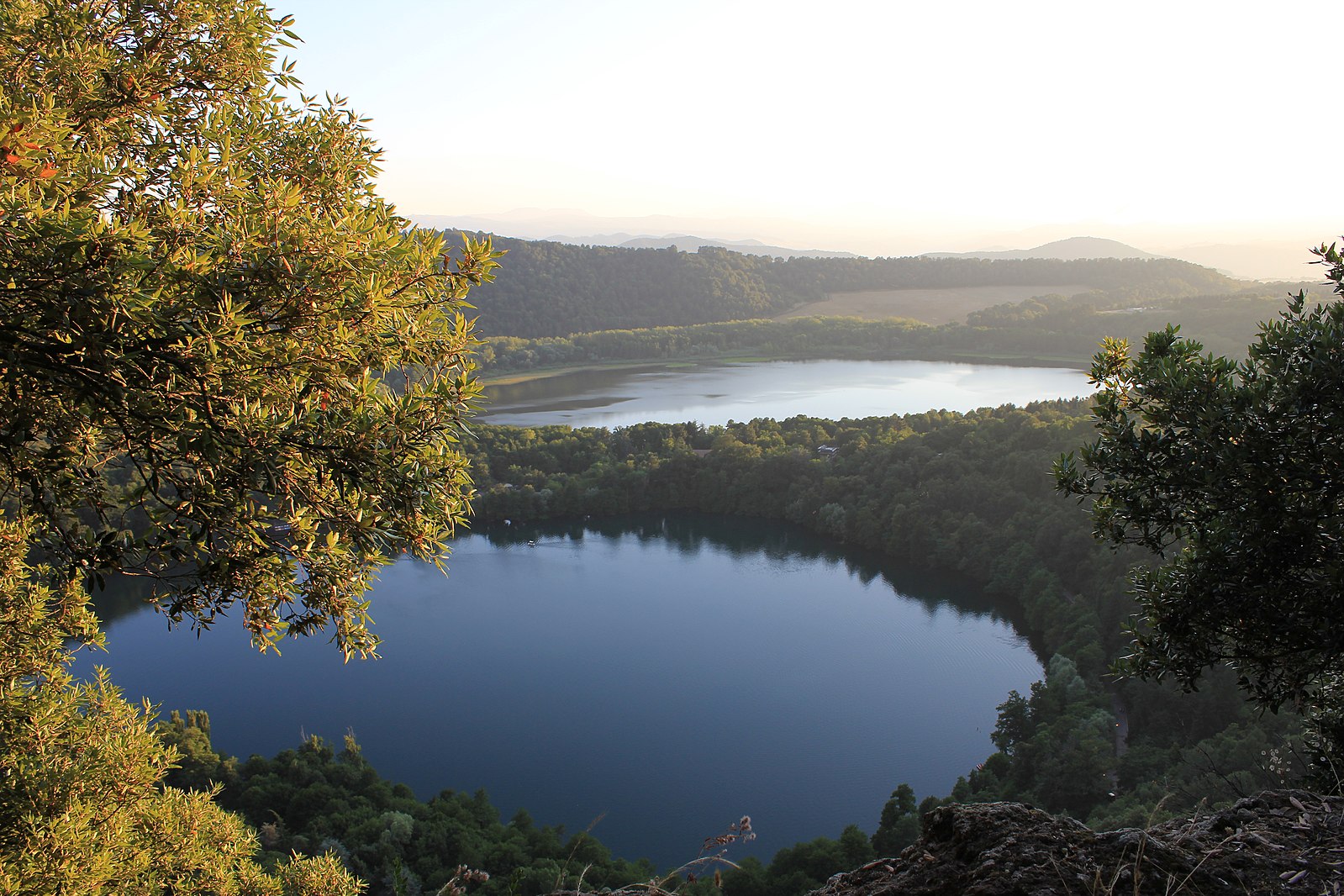 Vista di due laghi rotondi alberati, circondati da una fitta foresta, da un punto di osservazione più elevato. Il sole è basso, proiettando una luce calda sul paesaggio. I rami degli alberi incorniciano il primo piano.