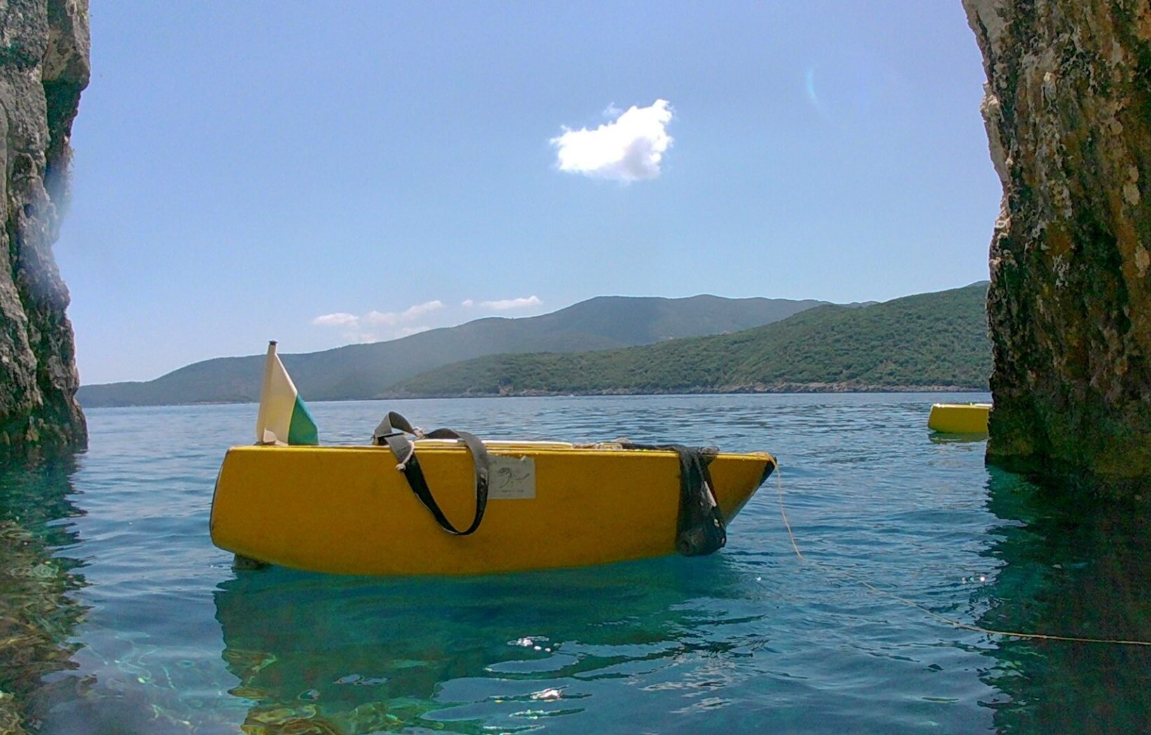 A bright yellow pedal boat with a small flag floats on calm blue water between two rocky cliffs, with green hills and a clear sky in the background.