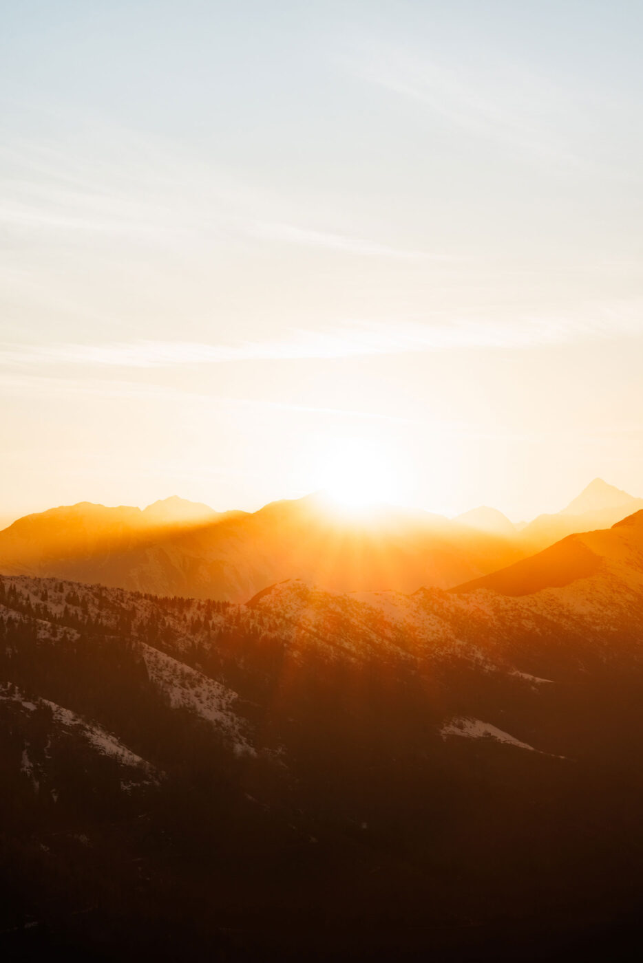 Il sole sorge o tramonta dietro una catena di montagne innevate. Il sole proietta un caldo bagliore dorato e tenui raggi sul cielo e sul paesaggio, illuminando le cime innevate e creando un'atmosfera tranquilla.