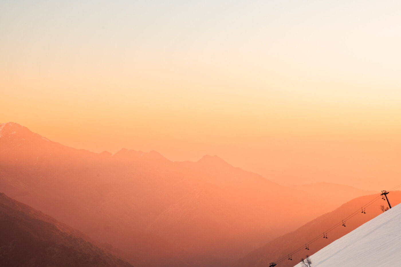 Alba in montagna nebbiosa - Paesaggio montano nebbioso all'alba o al tramonto, con tenui toni arancioni e rosa nel cielo e sagome nebbiose delle creste montuose. Uno skilift è visibile sul pendio destro.
