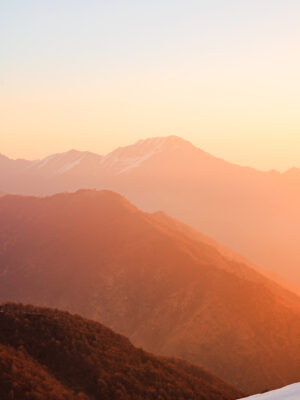 A mountain range bathed in soft, golden light during sunrise or sunset, with hazy layers of hills and a snow-capped peak in the distance under a clear sky.