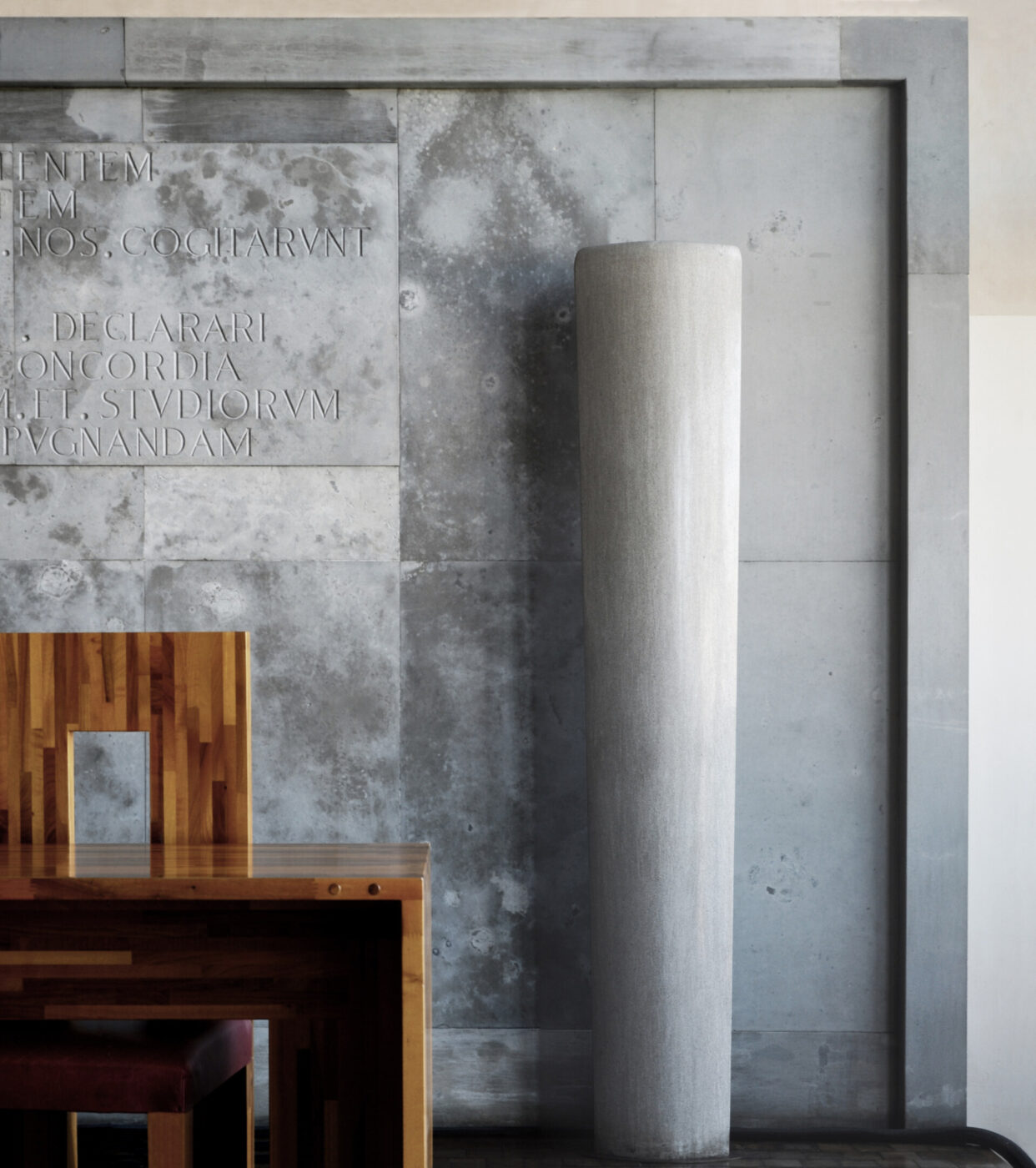 Design d'interni minimalista - Interni minimalisti con scrivania in legno con menù, un'alta colonna cilindrica e un muro in cemento con testo latino inciso. L'ambiente ha un'atmosfera moderna e sofisticata.