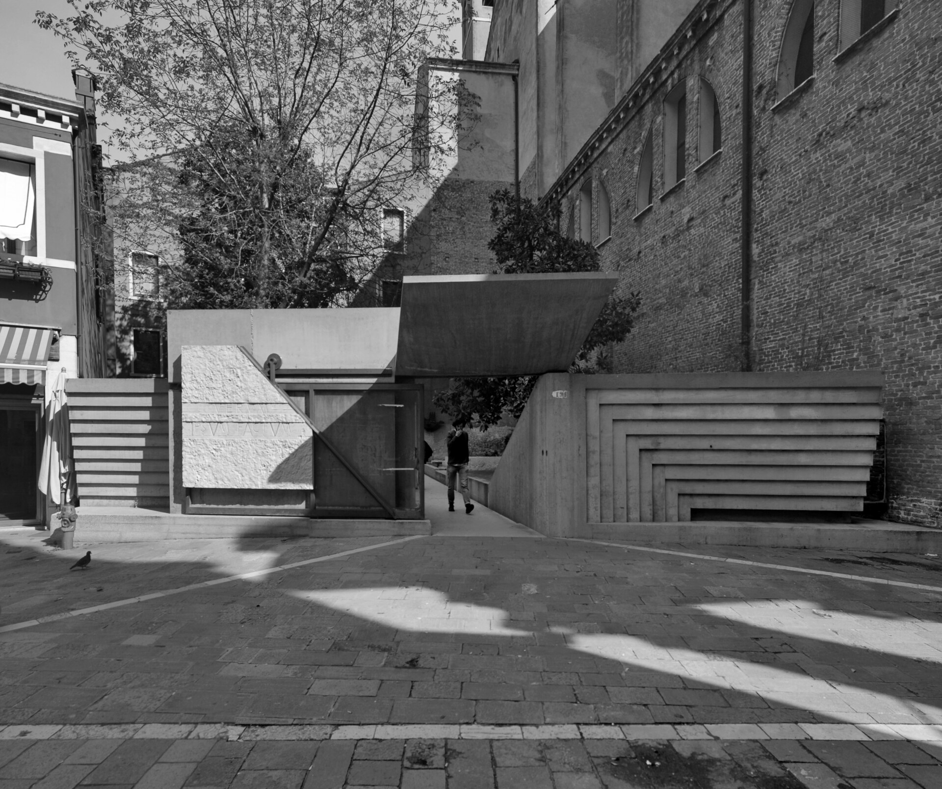 [Ingresso a uno spazio moderno] - Immagine in bianco e nero di un ingresso moderno e geometrico con elementi in cemento e legno. Gradini e pareti creano motivi a strati. Una persona entra dalla porta e un piccione è sul selciato.