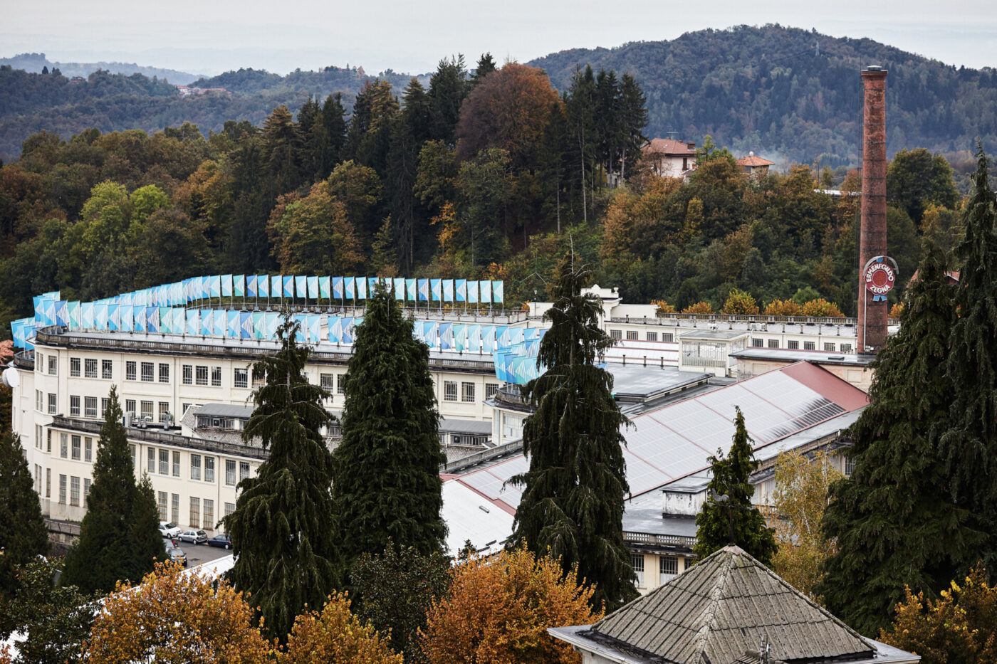 [Nessun titolo dell'immagine presente] - Un grande edificio industriale con pareti bianche e tetti bluastri, circondato da alti alberi sempreverdi e decidui, con colline e cielo coperto sullo sfondo. Un'alta ciminiera in mattoni si erge sulla destra.